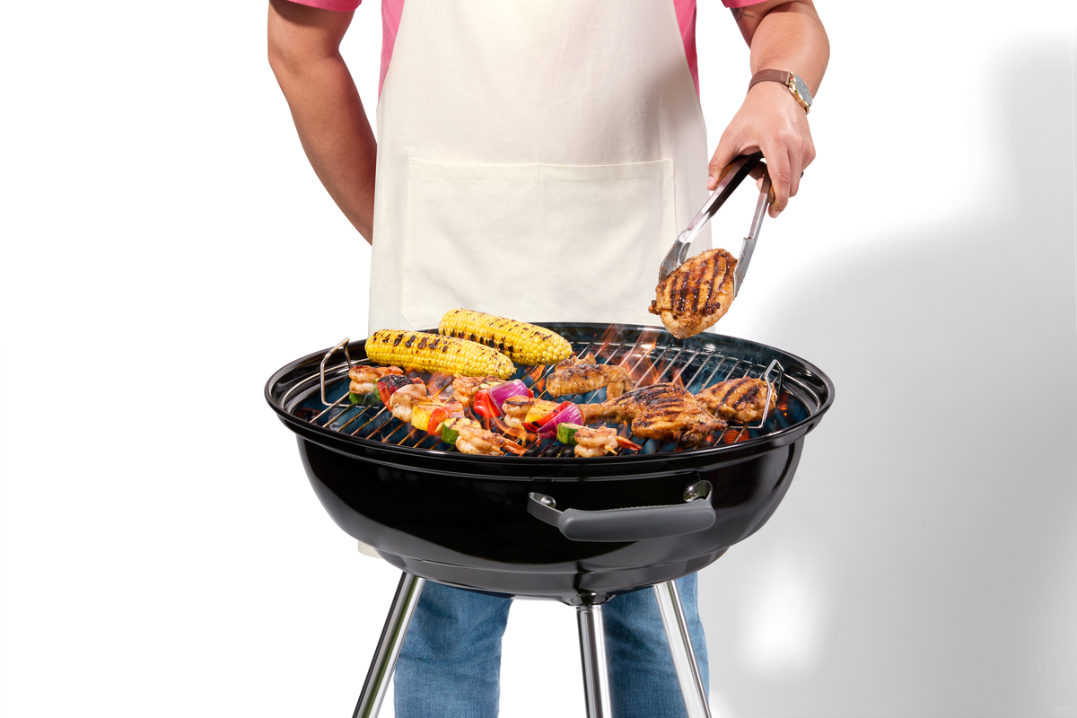 Man in a white apron grilling chicken, corn on the cob, and kebabs on a kettle-style grill.