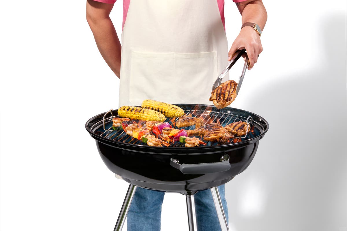 Man in a white apron grilling chicken, corn on the cob, and kebabs on a kettle-style grill.