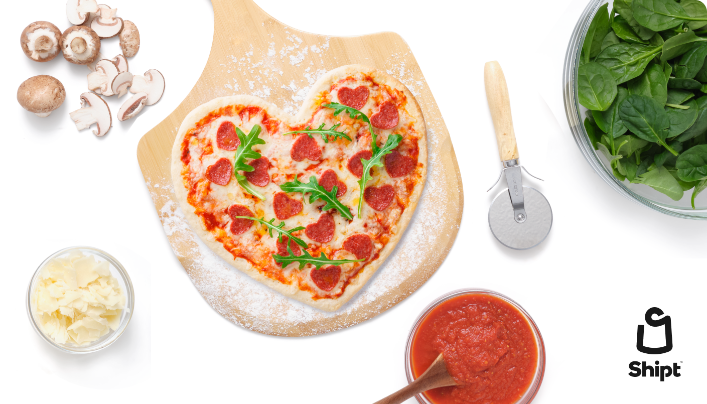 Pizza shaped like a heart on pizza stone, surrounded by various toppings like sauce, cheese, mushrooms, spinach