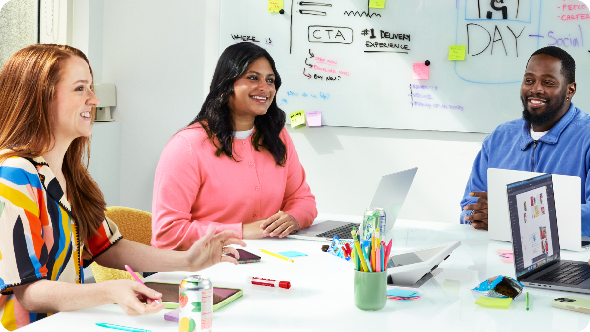 Three colleagues, two women and one man, are smiling and collaborating around a white conference table in a bright office setting.