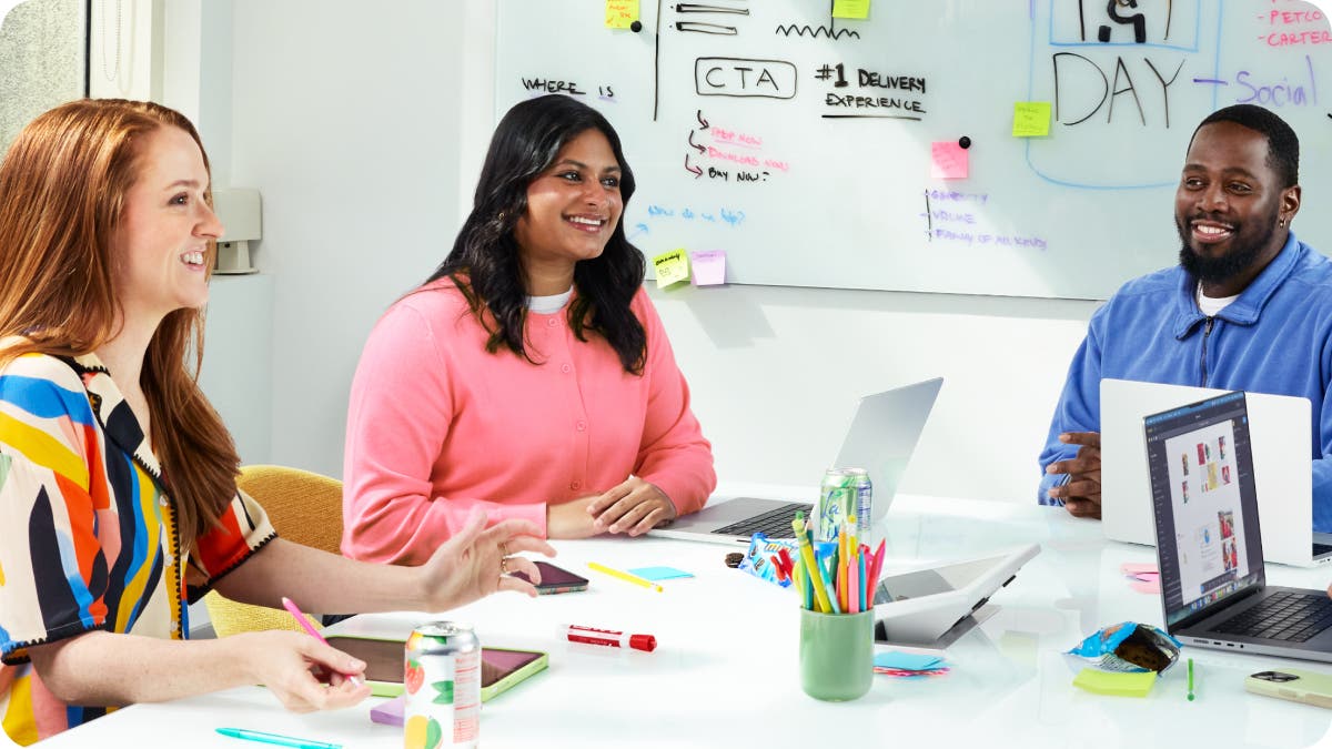 Three colleagues, two women and one man, are smiling and collaborating around a white conference table in a bright office setting.