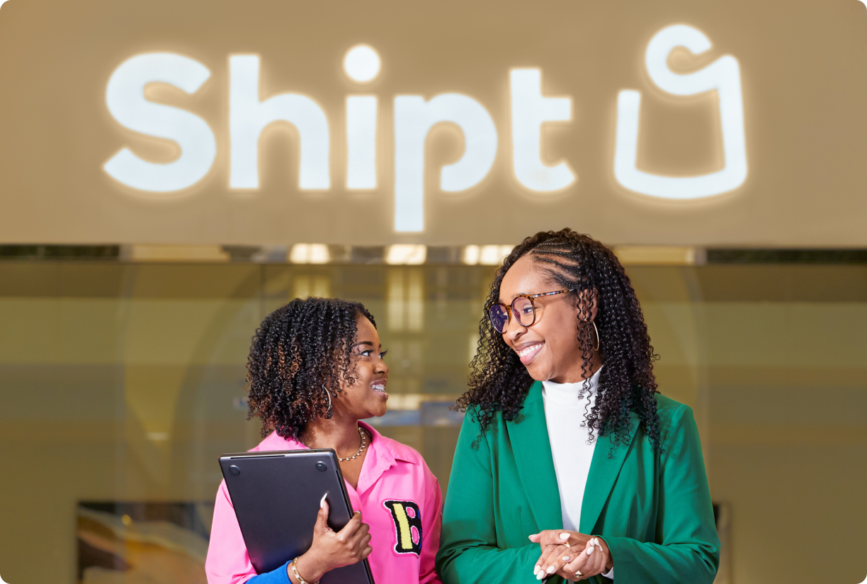 Two corporate employees talking in an office in front of a lighted Shipt sign.