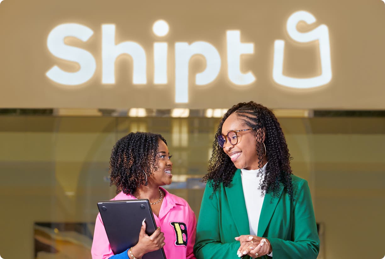 Two corporate employees talking in an office in front of a lighted Shipt sign.