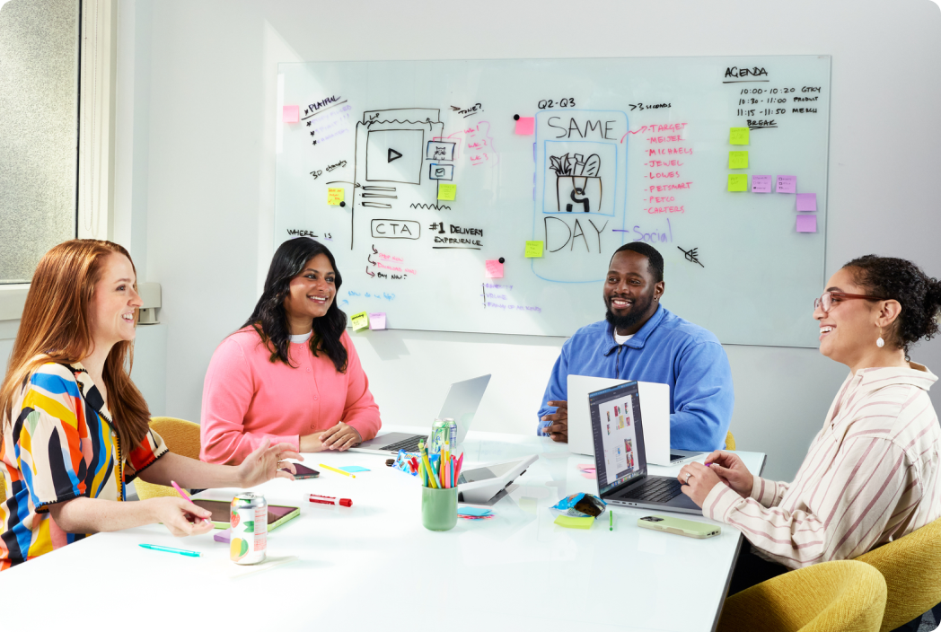 Four colleagues collaborating at a conference table with laptops in a bright office.