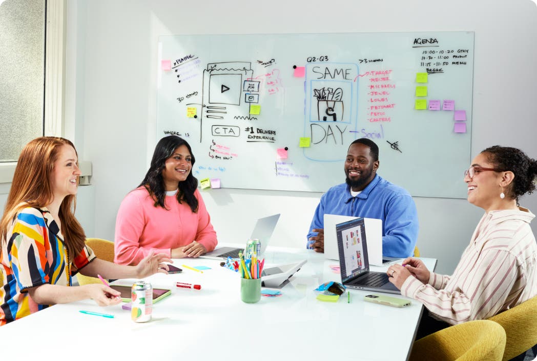 Four colleagues collaborating at a conference table with laptops in a bright office.