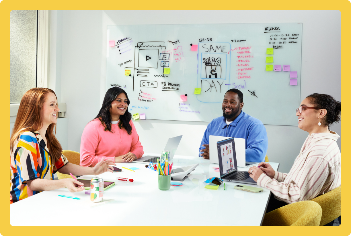 Four colleagues collaborating at a conference table with laptops in a bright office with yellow boarder.