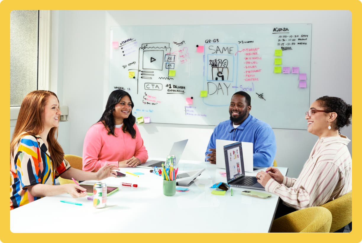 Four colleagues collaborating at a conference table with laptops in a bright office with yellow boarder.